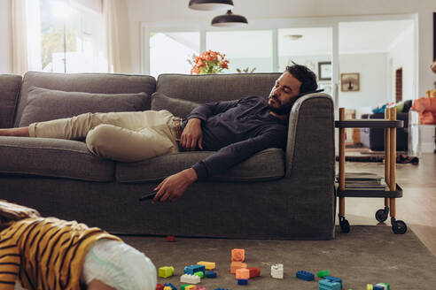 Mann entspannt sich auf der Couch mit seinem Kind, das auf dem Boden spielt. Kind spielt auf dem Boden mit Bauklötzen, während der Vater auf der Couch schläft. - JLPSF17027