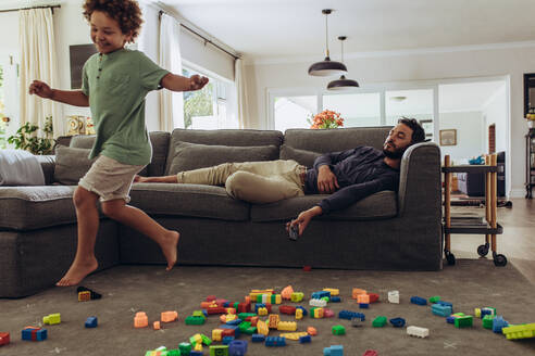 Ein Mann schläft auf der Couch, während sein Kind zu Hause spielt. Ein Junge rennt zu Hause mit Bauklötzen auf dem Boden herum. - JLPSF17028
