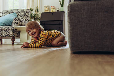 Lächelndes Kind sitzt zu Hause auf dem Boden. Fröhliches Kind versteckt sich hinter einer Couch und spielt Verstecken. - JLPSF17029