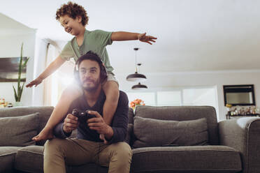 Man playing video game sitting on couch at home with his son sitting on his shoulders. Father and son having fun at home playing video game. - JLPSF17030