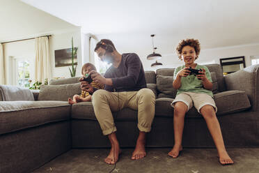Happy man having fun with his kids playing video game at home. Father playing video game sitting on couch with his two kids. - JLPSF17031