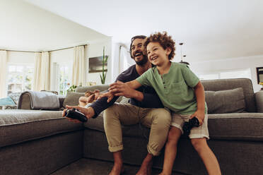 Father and son having fun playing video game at home. Man playing video game with son sitting on couch at home holding joysticks. - JLPSF17033