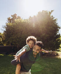 Ein Mann geht an einem sonnigen Tag im Park spazieren und trägt seinen Sohn auf dem Rücken. Ein glückliches Kind genießt die Fahrt auf dem Rücken seines Vaters mit offenen Armen. - JLPSF17035