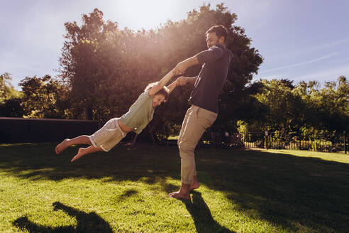 Vater schaukelt seinen Sohn herum und hält seine Hände, während er in einem Park steht. Vater und Sohn spielen an einem sonnigen Tag in einem Park. - JLPSF17039