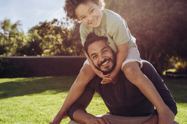 Smiling father and son sitting in a park on a sunny day. Boy sitting on the shoulders of his father at a park. - JLPSF17041