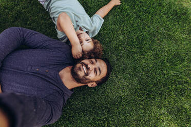 Man lying on grass in a park with his son. Top view of father and son lying on ground at a park with heads together. - JLPSF17043