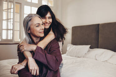 Ältere Frauen sitzen auf dem Bett mit ihrer Tochter, die ihre Hände hält. Lächelnde Mutter und Tochter sitzen zu Hause und verbringen Zeit miteinander. - JLPSF17058