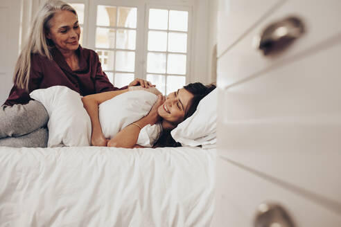 Lächelnde Frau, die auf einem Bett schläft und ein Kissen hält, während ihre Mutter neben ihr sitzt. - JLPSF17059