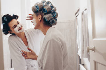 Mutter und Tochter in Bademänteln, die sich gemeinsam im Badezimmer pflegen. Zwei lächelnde Frauen mit Lockenwicklern auf dem Kopf, die Gesichtscreme auftragen. - JLPSF17064
