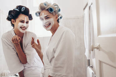 Frauen mit Lockenwicklern in den Haaren, die Gesichtscreme auftragen, stehen im Badezimmer. Glückliche Mutter und Tochter, die Spaß an der Pflege ihres Gesichts und ihrer Haare haben. - JLPSF17065