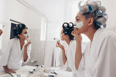 Frauen mit Lockenwicklern bei der Haarpflege zu Hause. Mutter und Tochter tragen im Badezimmer stehend Gesichtscreme auf - JLPSF17067