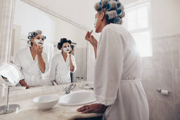 Mutter und Tochter in Bademänteln, die sich gemeinsam im Badezimmer pflegen. Zwei Frauen mit Lockenwicklern im Haar, die ihr Gesicht eincremen. - JLPSF17068