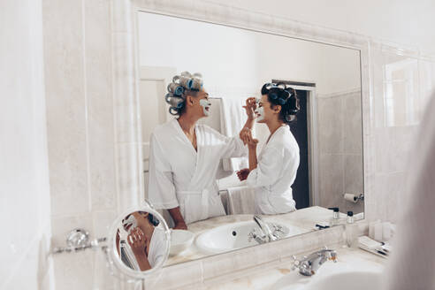 Frauen in Bademänteln und mit Lockenwicklern an den Haaren bei der Hauspflege. Ältere Frau cremt das Gesicht ihrer Tochter ein, die vor dem Spiegel steht. - JLPSF17069