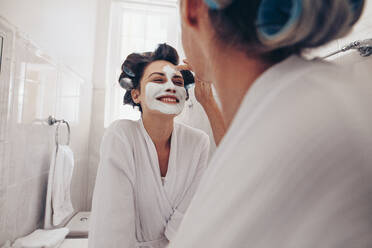 Lächelnde Frau im Bademantel, die sich zu Hause eine Gesichtsbehandlung gönnt. Zwei Frauen stehen im Badezimmer und tragen mit Lockenwicklern im Haar Gesichtscreme auf. - JLPSF17070