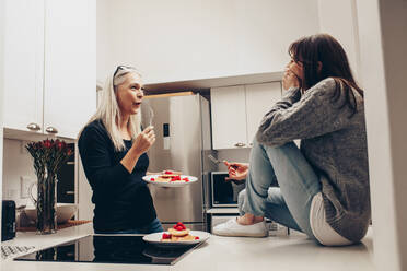 Ältere Frau isst Kekse von einem Teller, der in der Küche steht und sich mit einer Frau unterhält. Zwei Frauen unterhalten sich beim Essen von Snacks in der Küche. - JLPSF17082