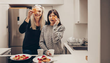 Glückliche Mutter und Tochter stehen in der Küche mit Tellern voller Kekse. Zwei lächelnde Frauen in der Küche halten sich Kekse vor die Augen. - JLPSF17083