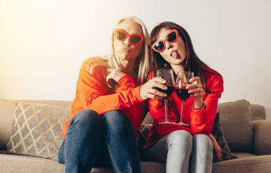 Women in similar outfit and fancy eyewear toasting wine and making faces sitting on couch. Mother and daughter having fun sitting at home holding wine glasses. - JLPSF17086