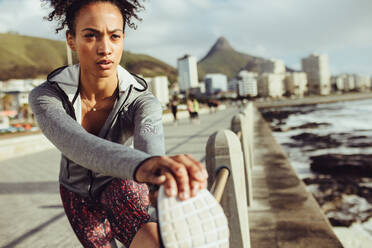 Fitte junge Läuferin beim Training im Freien. Frau in Sportkleidung streckt ihre Beine auf einem Geländer an der Strandpromenade. - JLPSF17093