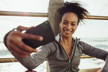 Lächelnde junge Frau in Sportkleidung, die ein Selfie mit ihrem Mobiltelefon macht. Eine Frau entspannt sich und macht ein Selfie nach dem Training am Meer. - JLPSF17096