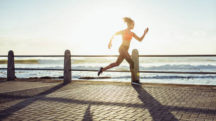 Fitte junge Frau, die schnell über eine Strandpromenade rennt. Seitenansicht einer Läuferin, die bei Sonnenuntergang sprintet. - JLPSF17102