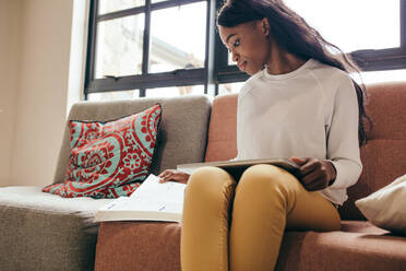 African girl sitting on a sofa with a tablet pc reading a book. Young student studying indoors at college campus. - JLPSF17104