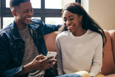 Zwei afroamerikanische College-Studenten benutzen ein Handy auf dem Campus. Ein glücklicher junger Mann und eine Frau sitzen auf dem Sofa mit einem Smartphone. - JLPSF17115