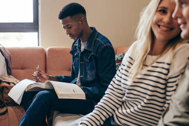 Afrikanischer männlicher Student, der sich Notizen macht, während er auf einem Sofa auf dem College-Campus sitzt und Bücher liest. - JLPSF17123