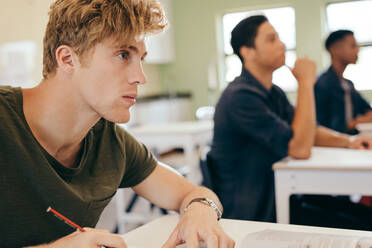 Männlicher Student, der im Klassenzimmer sitzend der Vorlesung zuhört, mit anderen Studenten im Hintergrund. - JLPSF17145