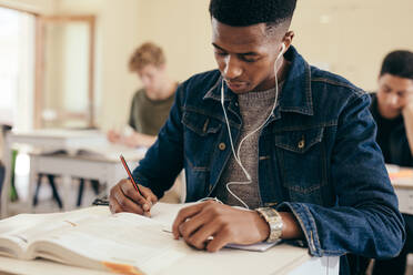 Teenager, der in einem Klassenzimmer der High School sitzt und in ein Buch schreibt. Männlicher Student mit Kopfhörern, der während einer Vorlesung in einem College-Klassenzimmer Notizen macht. - JLPSF17148