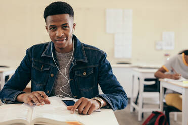 Afrikanischer Teenager sitzt in einem Klassenzimmer mit Büchern. Männlicher Student mit Kopfhörern sitzt in einem College-Klassenzimmer. - JLPSF17149