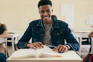 Lächelnder männlicher Student in einem College-Klassenzimmer mit Büchern. Teenager-Junge sitzt in einem College-Klassenzimmer. - JLPSF17150