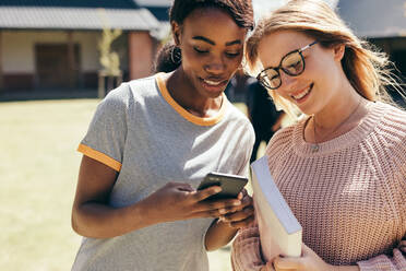 Zwei junge Frauen auf dem College-Campus, die auf ihr Handy schauen. Highschool-Mädchen, die auf dem Universitätscampus mit einem Smartphone spazieren gehen. - JLPSF17154