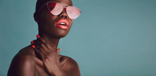 Stilvolles weibliches Modell mit lebhaftem Make-up und trendiger Sonnenbrille vor grauem Hintergrund. Glamouröse Frau mit rotem Lippenstift posiert im Studio. - JLPSF17179