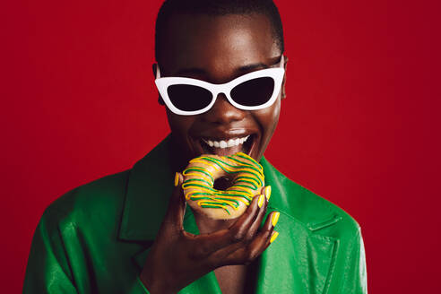Modische Frau isst Donut auf rotem Hintergrund. Fröhliches weibliches Model mit trendiger Sonnenbrille beißt in einen Donut. - JLPSF17213