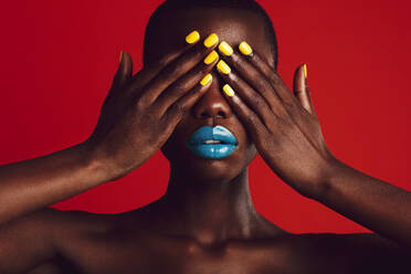 Nahaufnahme einer afrikanischen Frau mit buntem Make-up, die ihre Augen mit den Händen bedeckt. Weibliches Modell mit lebhaftem Make-up auf rotem Hintergrund. - JLPSF17217