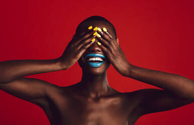Fröhliche afrikanische Frau mit lebhaftem Make-up, die ihre Augen mit den Händen bedeckt. Weibliches Modell lächelnd auf rotem Hintergrund. - JLPSF17218