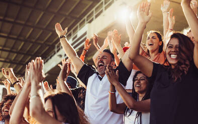 Eine Gruppe von Sportfans jubelt während eines Spiels im Stadion. Aufgeregte Menschen stehen mit erhobenen Armen da, klatschen und schreien, um ihre Mannschaft anzufeuern. - JLPSF17225
