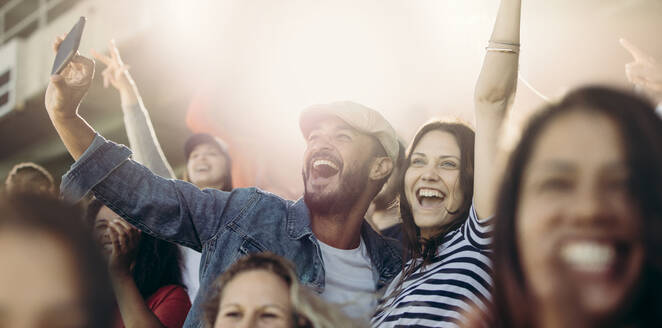 Aufgeregtes Paar, das jubelt und ein Selfie macht, während es im Stadion ist. Fröhliche Fußballfans machen ein Selfie, während sie ein Spiel im Stadion sehen. - JLPSF17273
