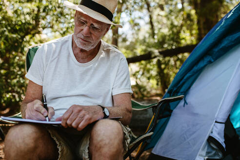Älterer Mann sitzt vor einem Zelt und schreibt in ein Buch. Älterer kaukasischer Mann schreibt ein Buch auf dem Campingplatz. - JLPSF17313