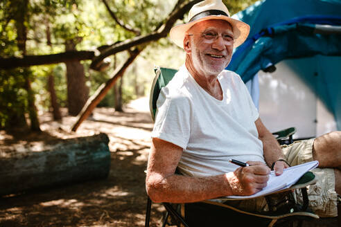 Smiling senior man sitting in front of a tent and writing in a book. Retired man looking at camera and smiling while writing a book at campsite. - JLPSF17314