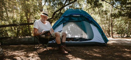 Älterer Mann schreibt ein Buch auf dem Campingplatz. Älterer männlicher Reisender schreibt ein Buch auf dem Campingplatz. Er sitzt auf einem Stuhl außerhalb des Zeltes. - JLPSF17315