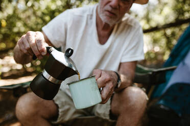 Nahaufnahme eines älteren Mannes, der auf einem Campingplatz Kaffee in einen Becher gießt. Männlicher Reisender, der auf einem Campingausflug Kaffee trinkt. - JLPSF17317