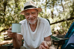Lächelnder älterer Mann mit erfrischendem Kaffee auf dem Campingplatz. Männlicher Reisender mit einer Tasse Kaffee in der Hand und Blick in die Kamera auf einem Campingausflug. - JLPSF17318
