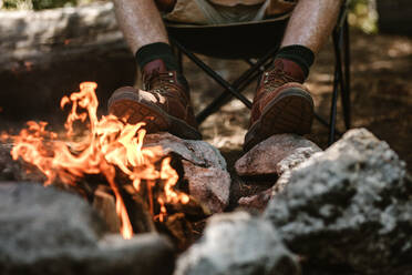 Mann in Stiefeln wärmt sich die Füße am Feuer auf dem Campingplatz. Ausschnitt eines älteren Mannes, der am Lagerfeuer sitzt. - JLPSF17319