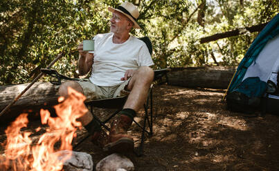 Ein Mann im Ruhestand sitzt am Lagerfeuer und trinkt Kaffee. Ein älterer Mann zeltet im Wald und trinkt einen erfrischenden Kaffee am Morgen. - JLPSF17320