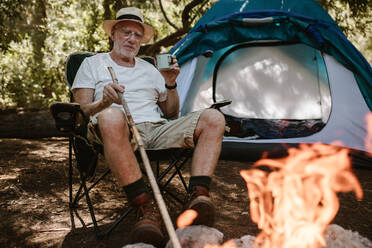 Rentner sitzt entspannt in der Nähe von Lagerfeuer und mit einer Tasse Kaffee. Senior Mann Camping im Wald mit einem erfrischenden Kaffee am Morgen. - JLPSF17322