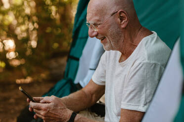 Lächelnder älterer Mann, der im Zelt sitzt und sein Handy benutzt. Rentner, der in der Natur zeltet und eine Textnachricht auf seinem Handy liest. - JLPSF17329