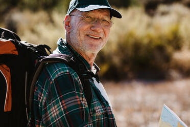 Senior man carrying a backpack looking at camera and smiling. Fit old man on a hiking trip. - JLPSF17334