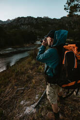 Älterer Mann auf Wandertour, der mit einer Digitalkamera fotografiert. Älterer Mann, der am Fluss steht und auf Wandertour Fotos macht. - JLPSF17353