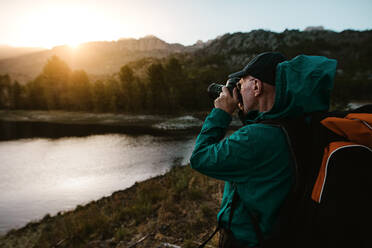 Alter Mann im Wanderurlaub, der die schöne Aussicht mit einer Digitalkamera fotografiert. Älterer Mann steht am Fluss im Wald und macht Fotos. - JLPSF17354